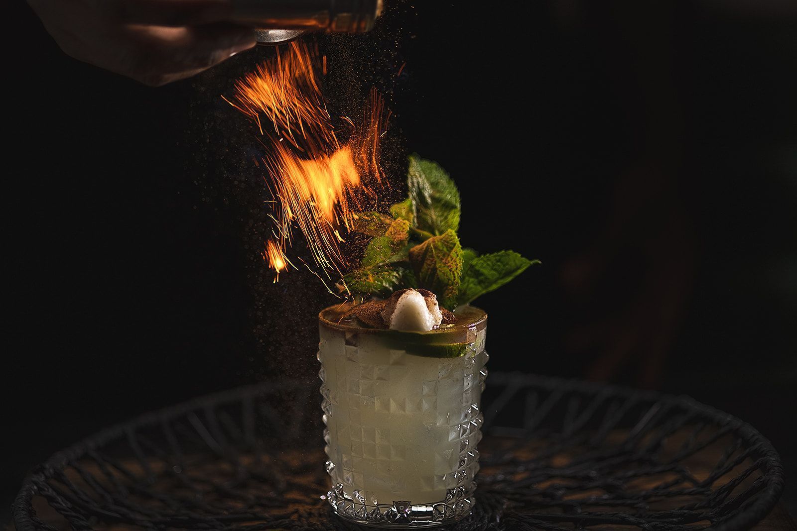 A Mexican-inspired cocktail garnished with mint leaves and lime, topped with a flame spark for a dramatic effect.