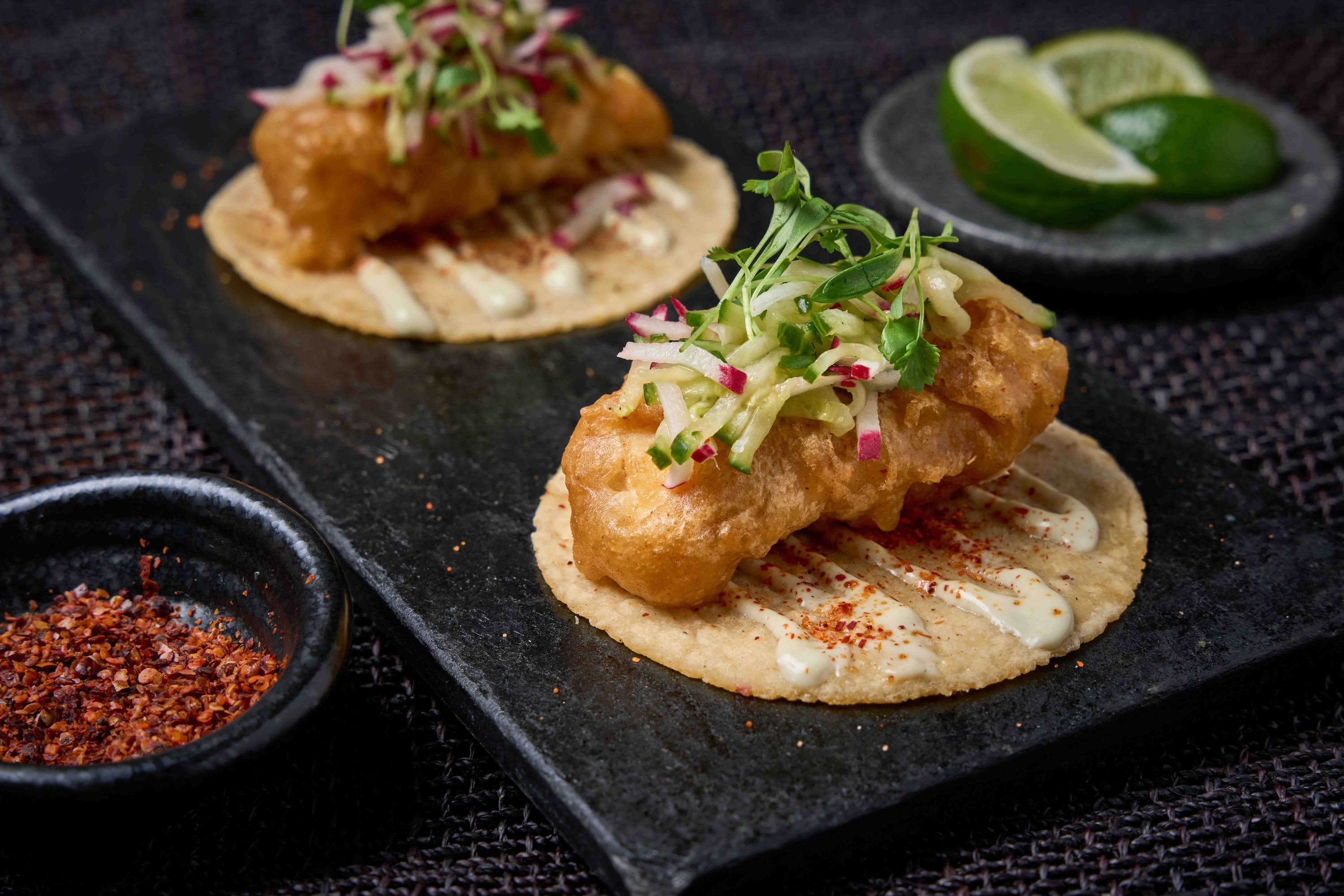Crispy fish tacos topped with slaw served on corn tortillas with lime wedges and chili powder on a black slate.