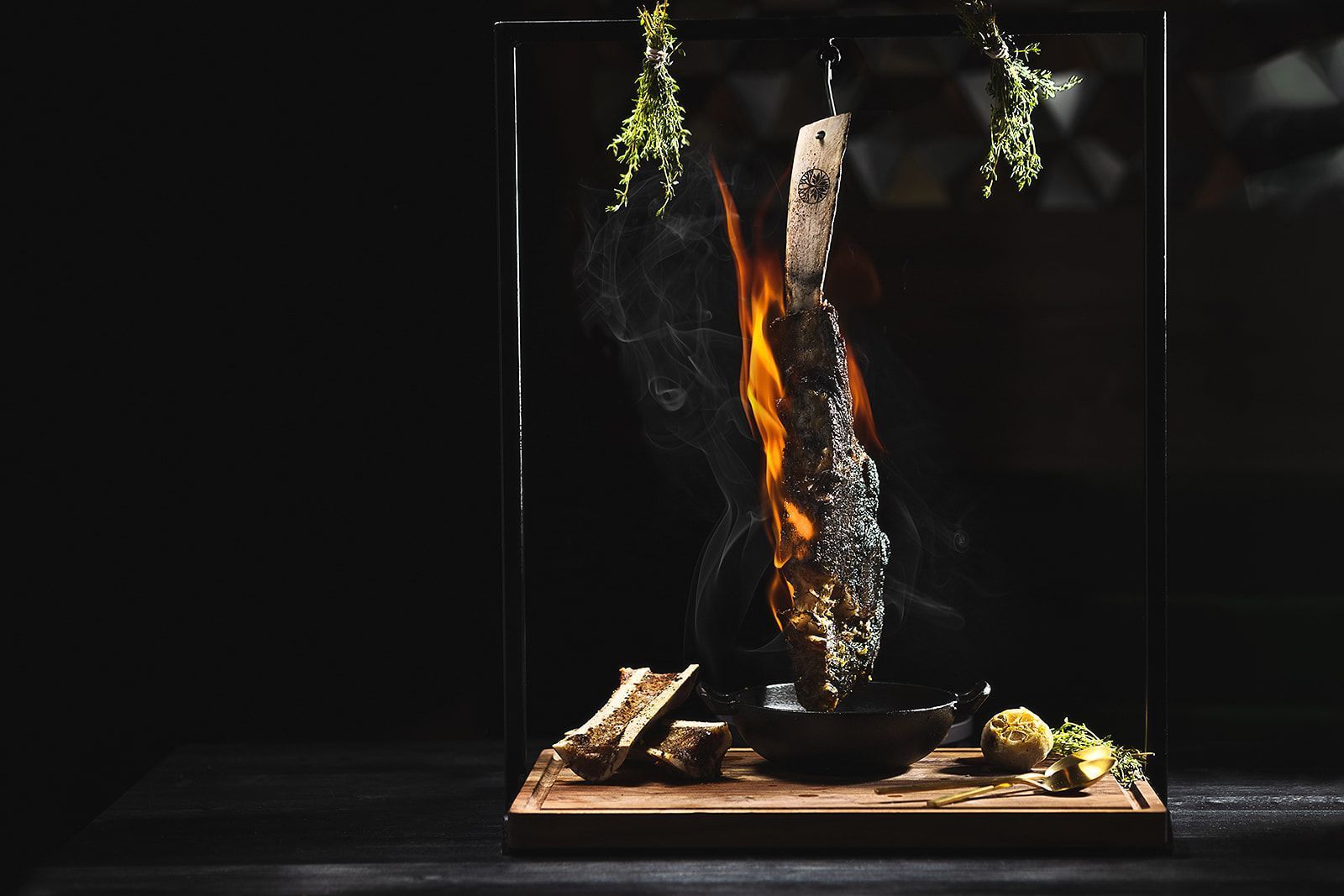A tomahawk steak hanging on a hook with flames and herbs, served with roasted garlic and herbs on a wooden board.