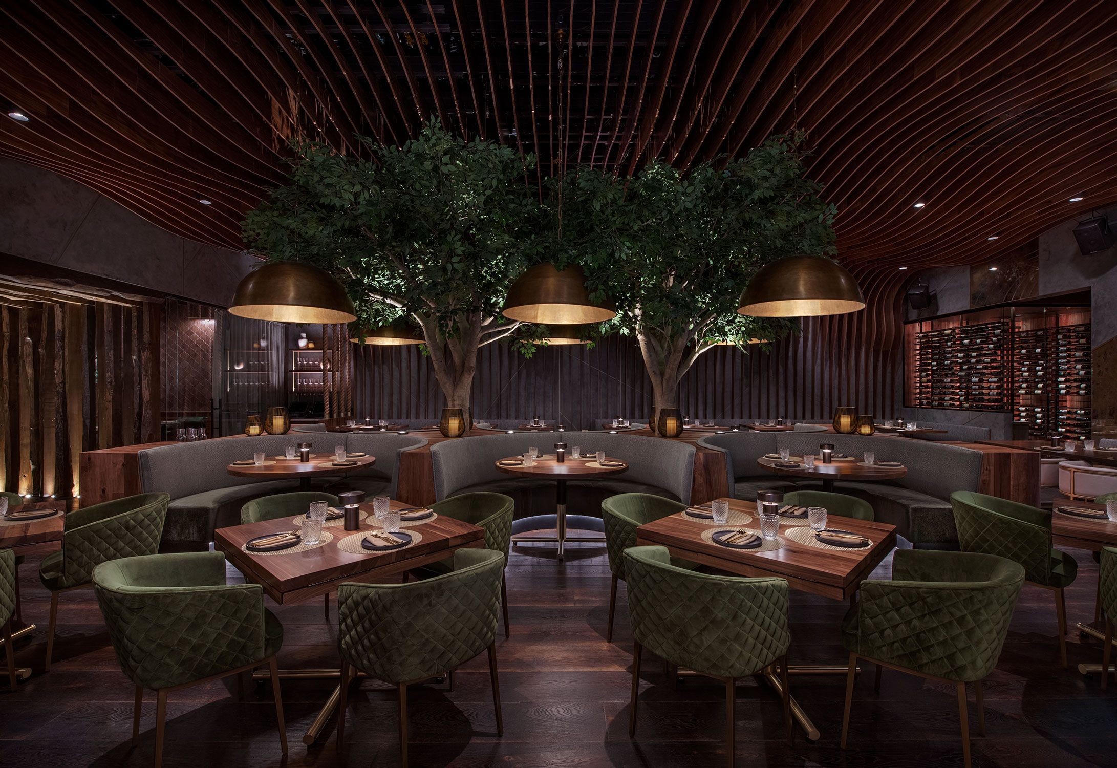 Upscale dining area with curved seating, green velvet chairs, wooden tables, large pendant lights, and indoor trees.