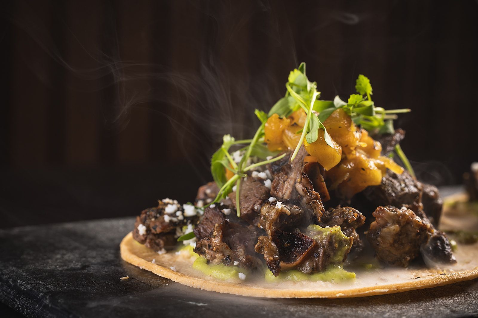 Mexican-inspired taco with grilled meat, garnished with greens and diced yellow peppers on a thin tortilla base.