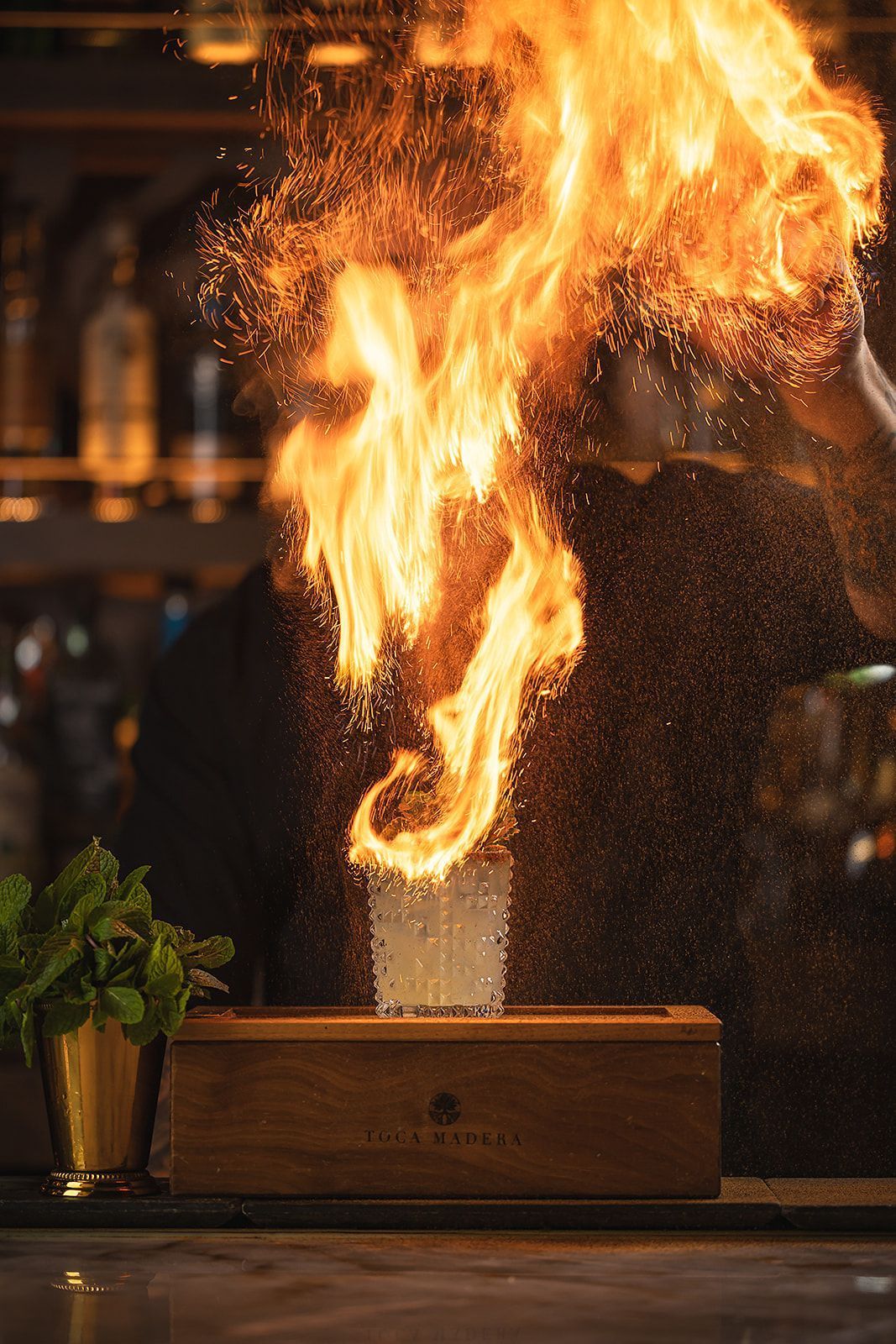 A flaming cocktail is served in a textured glass on a wooden board with the Toca Madera logo, garnished with fresh mint.