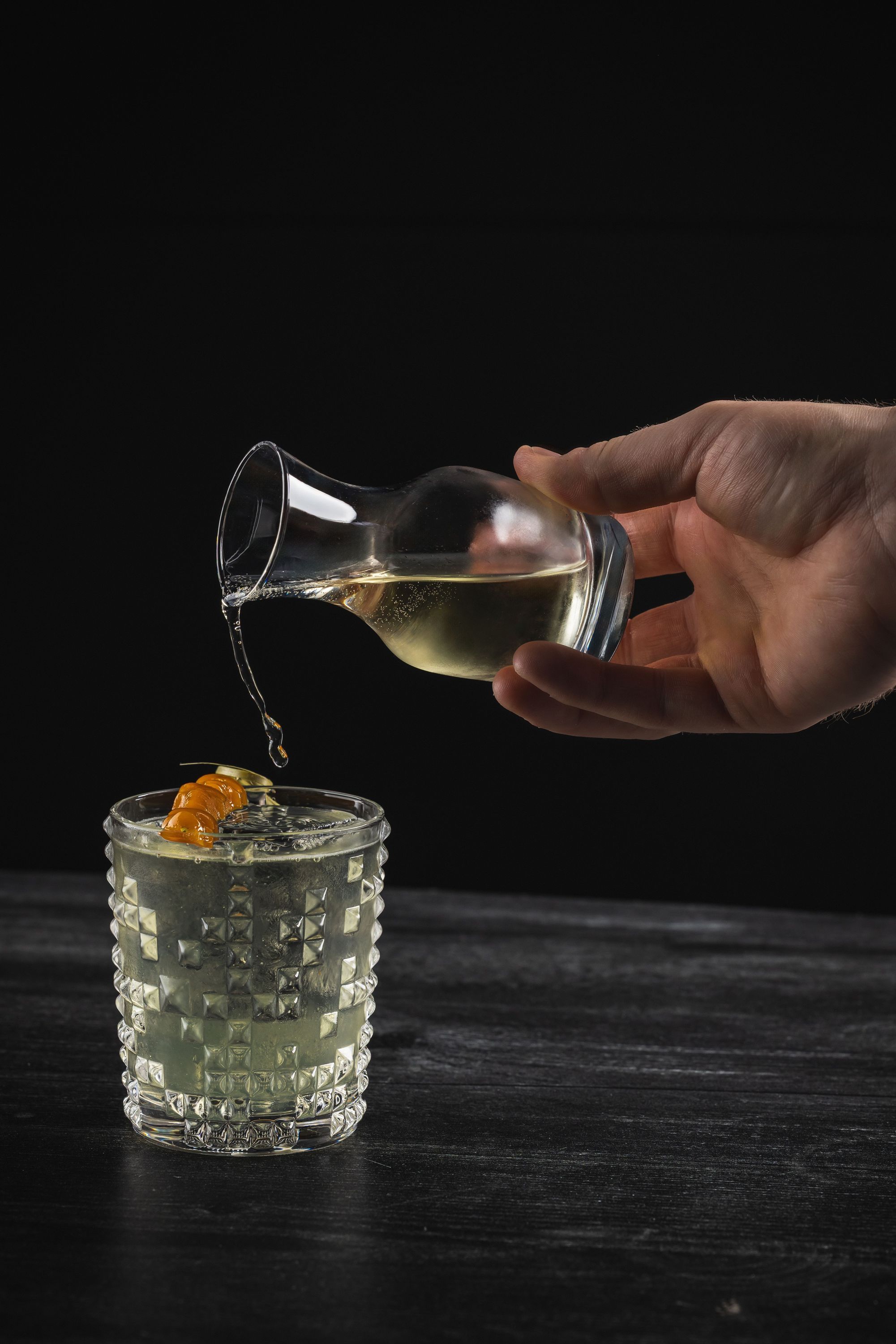 Hand pouring clear liquid from a small glass pitcher into a textured glass with a light beverage and orange peel garnish.
