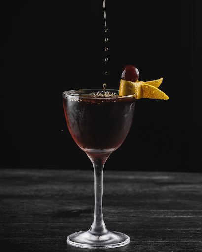 Dark cocktail being poured into a coupe glass garnished with an orange twist and a cherry on a dark background.
