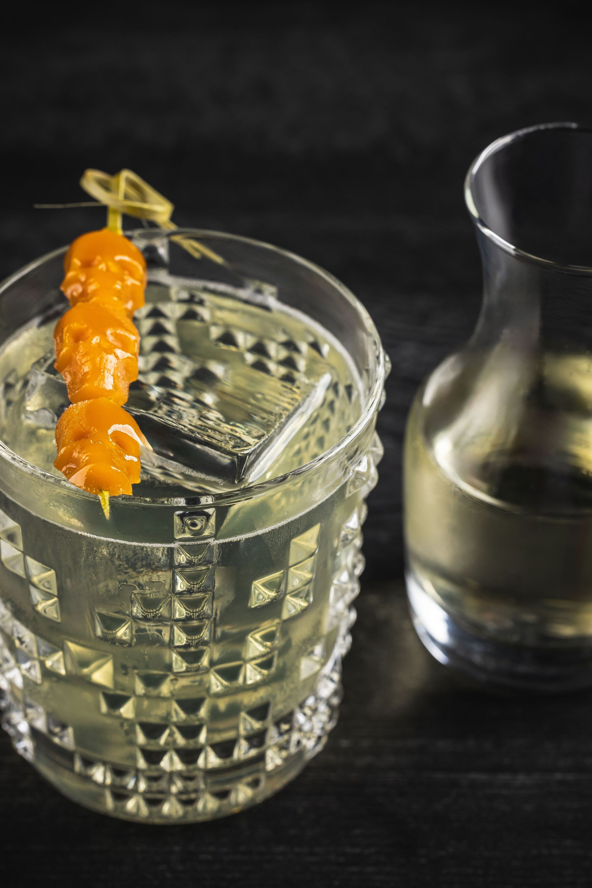 Clear cocktail with an orange garnish served in a textured glass alongside a carafe on a dark surface