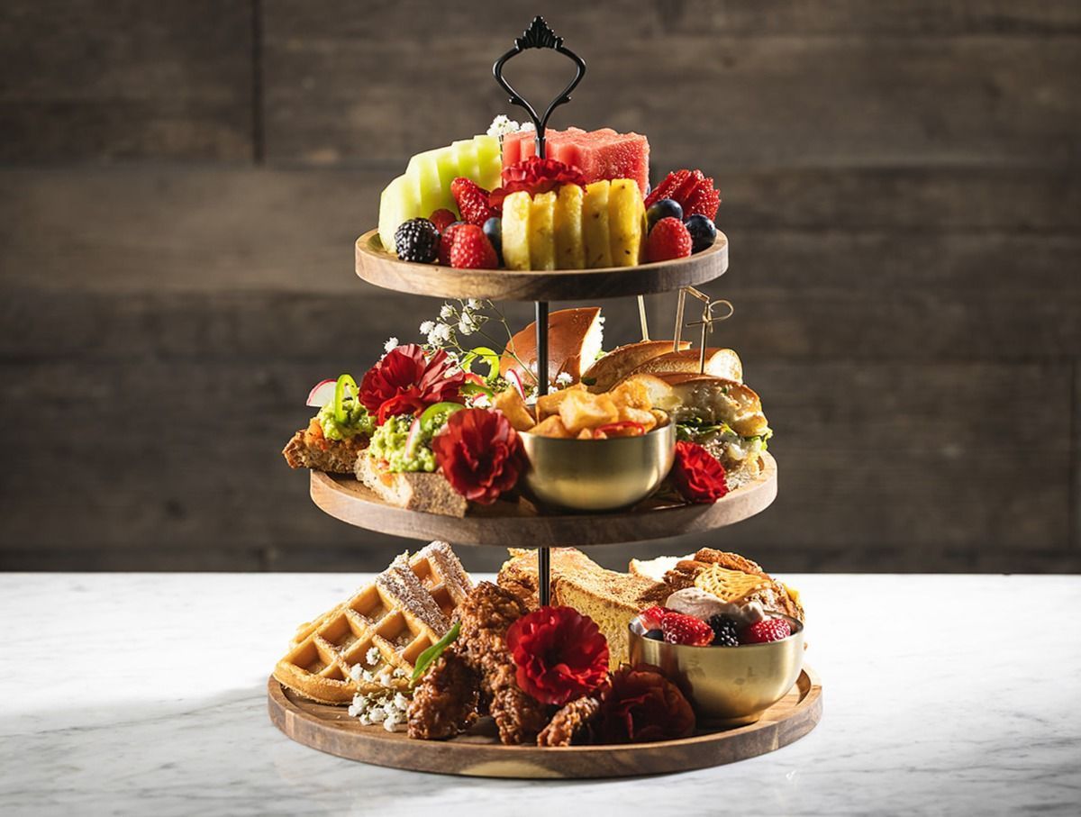 Three-tiered platter with assorted fresh fruit, sandwiches, fried chicken, waffles, and bowls of berries on a marble table.