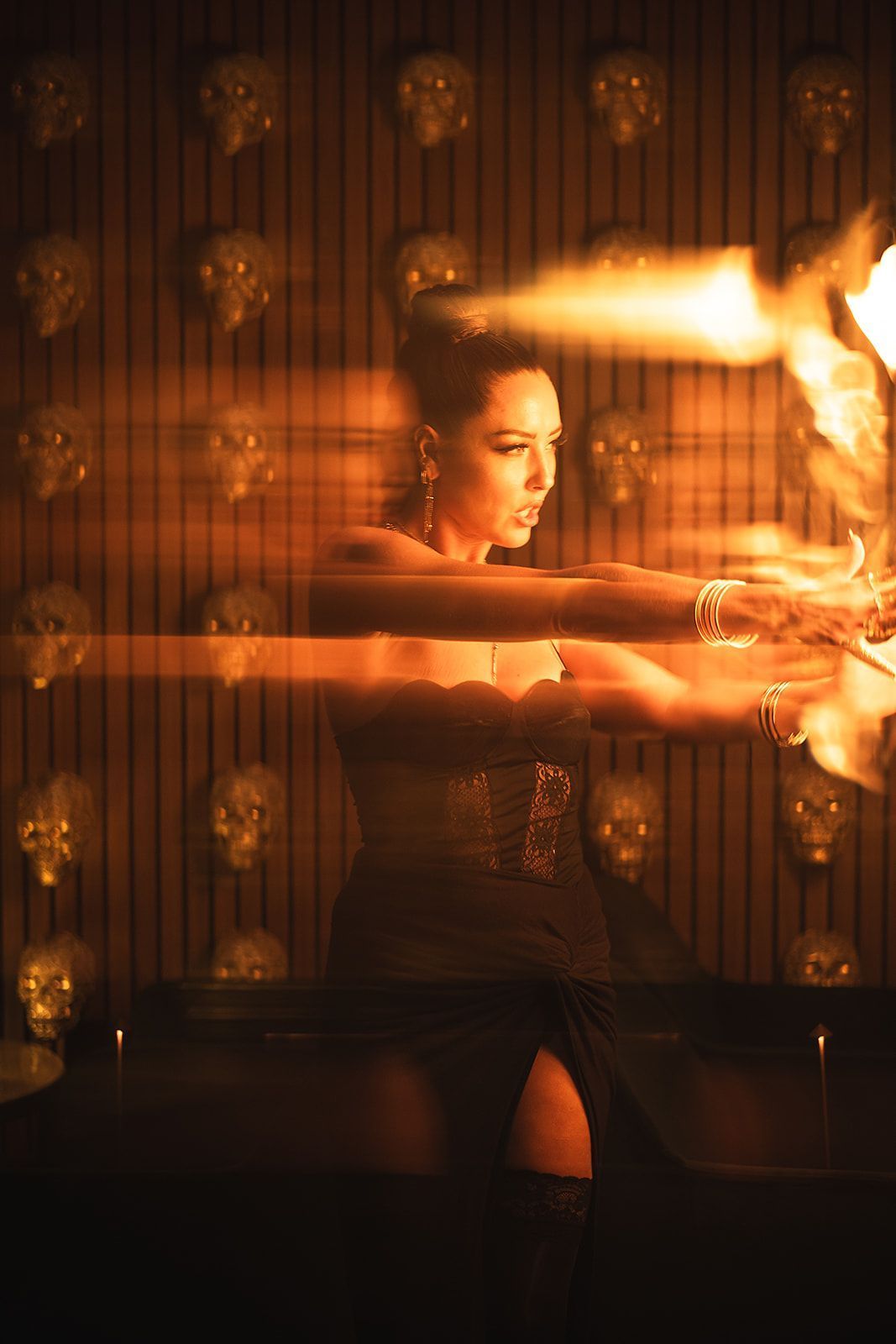 Performer in a black dress holding fire sticks in a dimly lit setting with skull decorations on the wall.