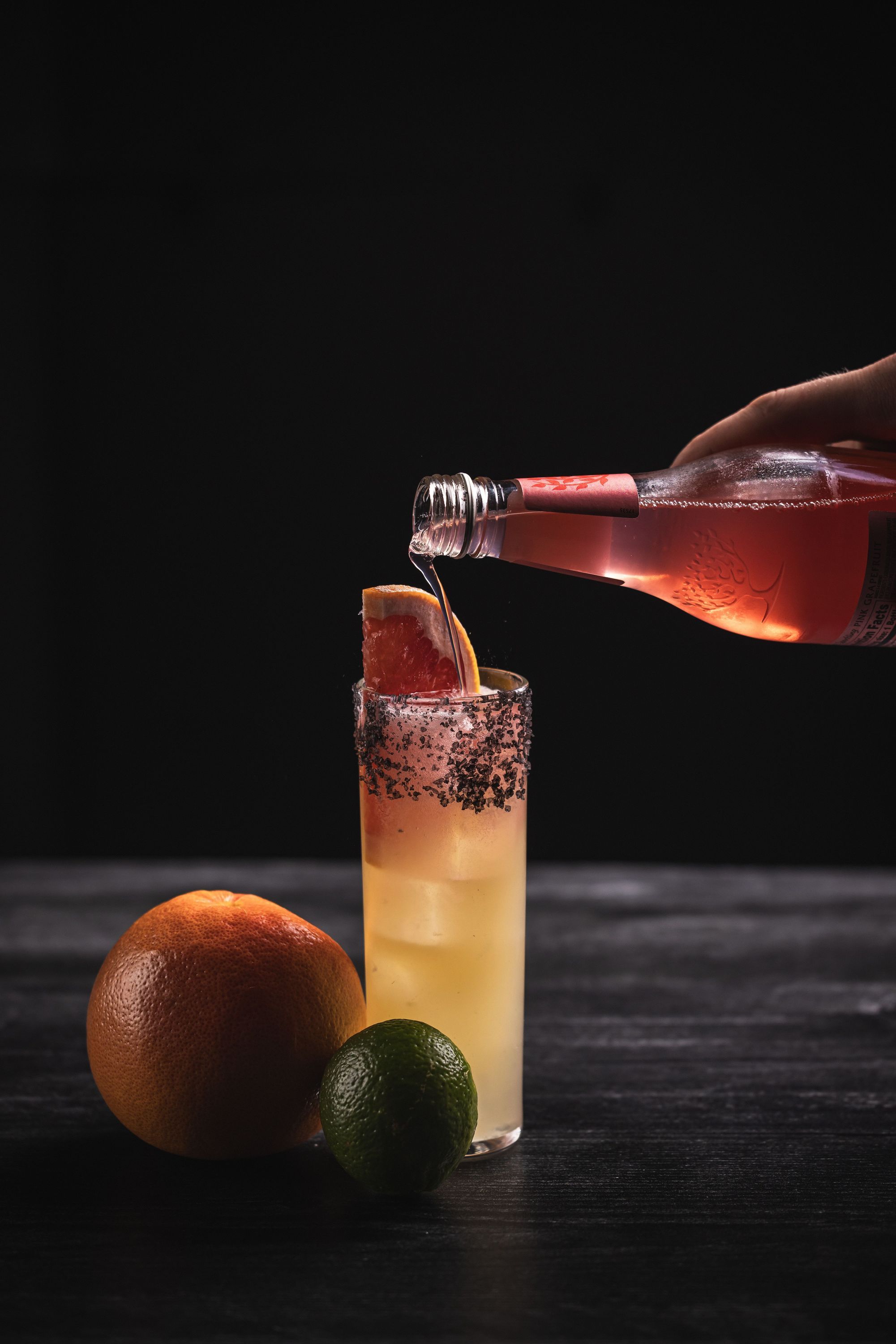 Cocktail with grapefruit slice and black salt rim next to lime and grapefruit, poured from a pink bottle at the restaurant.