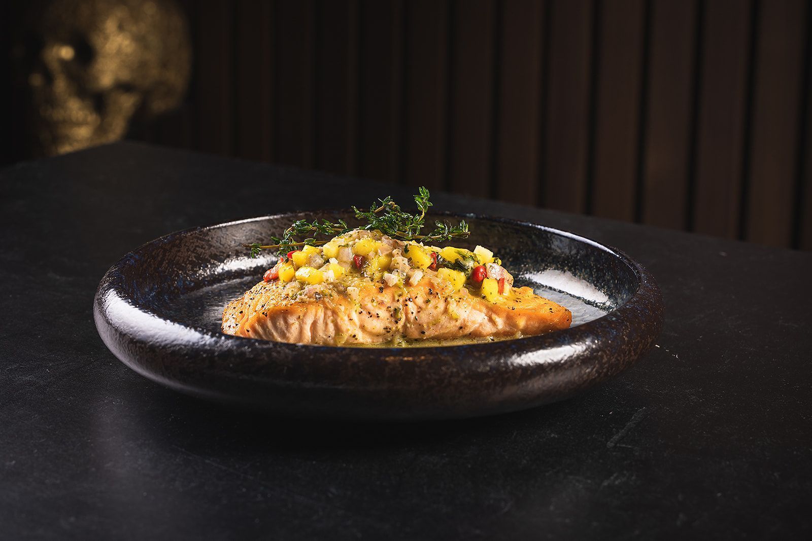 Seared salmon topped with a fresh mango salsa and garnished with herbs on a black ceramic plate, Toca Madera Las Vegas.