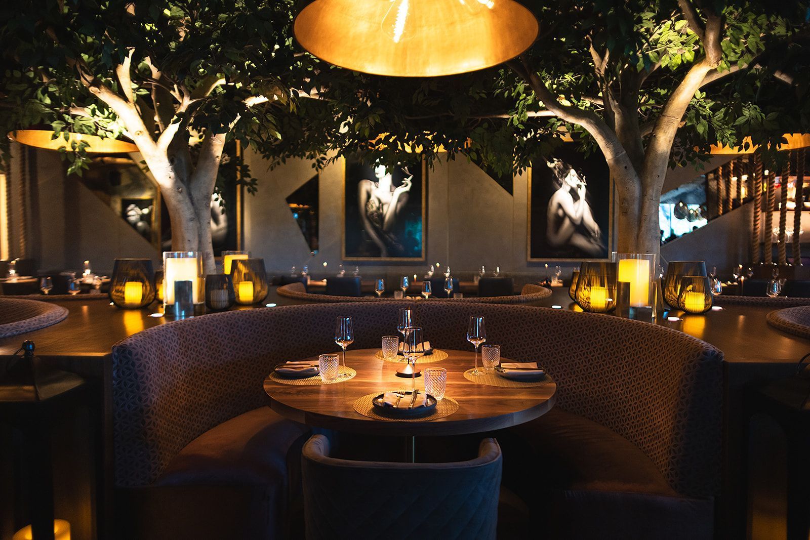 Round wooden table set with glassware and napkins surrounded by a curved booth and illuminated by warm pendant lights.