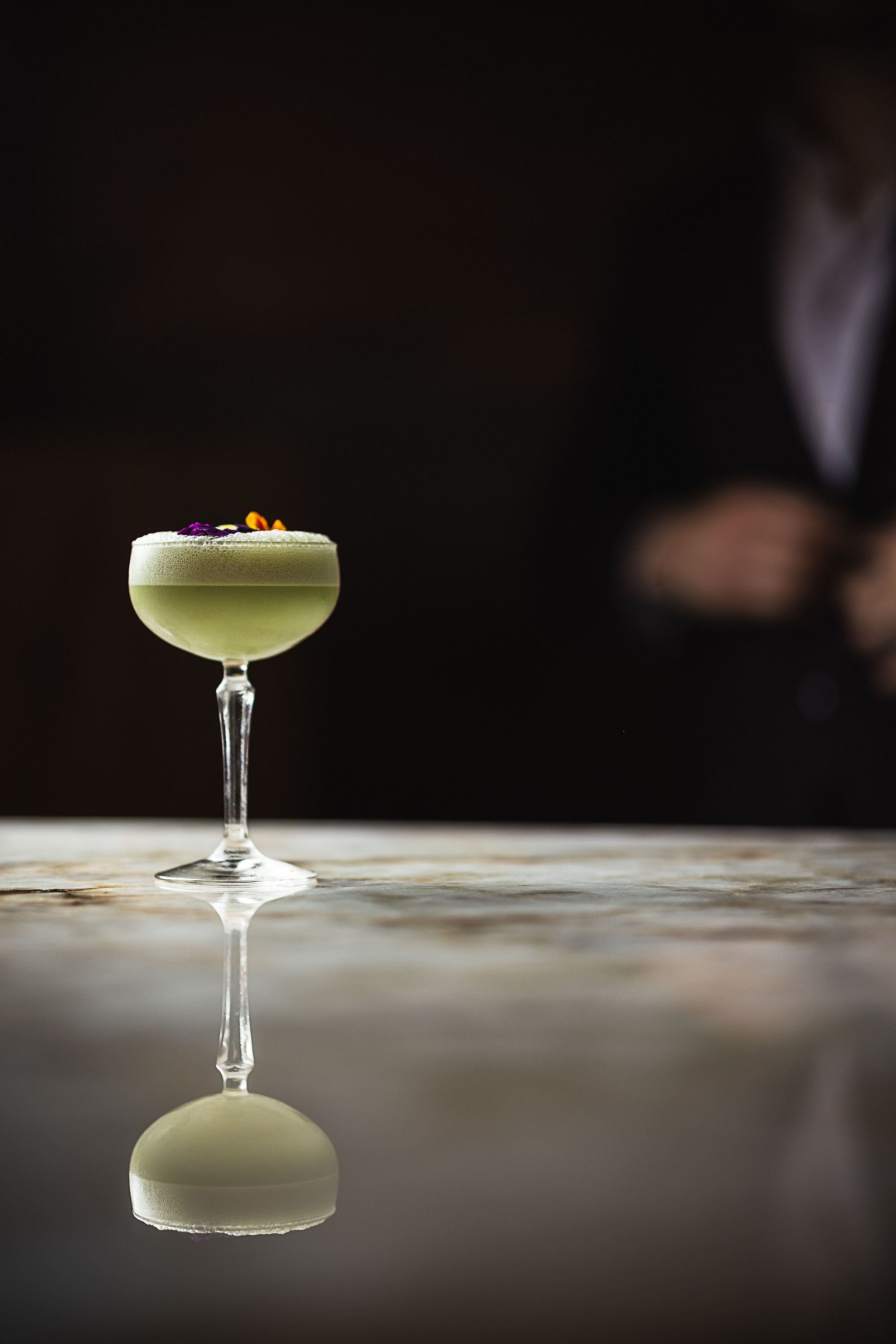 A delicate green cocktail with floral garnish in a coupe glass, set on a reflective marble bar surface.