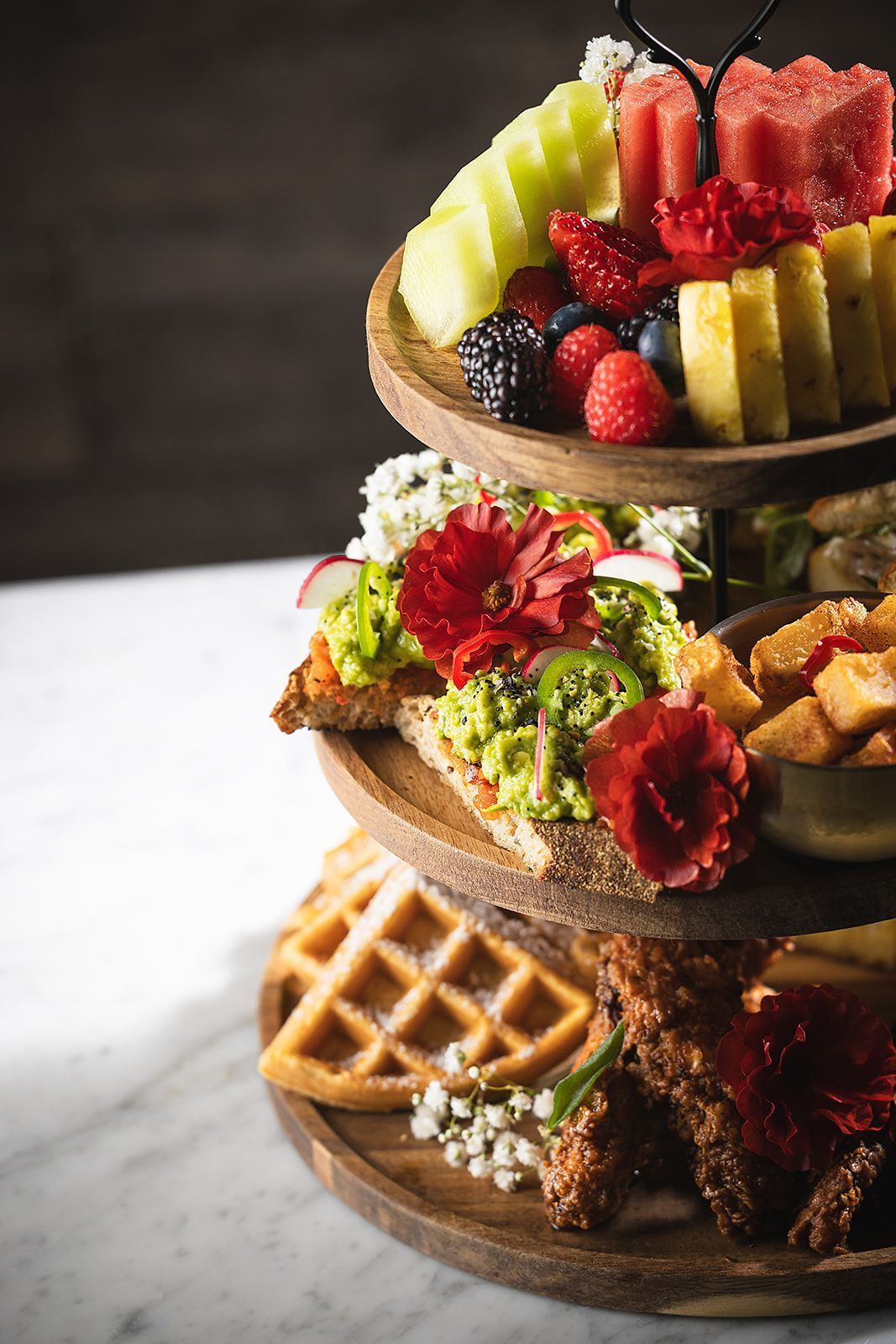 Three-tier wooden tray holding fresh fruits, avocado toast with flowers, fried chicken, and waffles on a marble table.